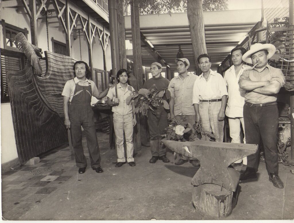 Fotografía en blanco y negro de un grupo de ocho hombres posando en un taller artístico. Al fondo se observa una escultura metálica de gran tamaño en forma de ave. A la izquierda, un hombre con overol sostiene una herramienta, posiblemente el artista. En el centro, algunos hombres sostienen hojas y flores. A la derecha, un hombre con sombrero y pipa cruza los brazos. En primer plano, se ve un yunque sobre un tronco de madera. La arquitectura del lugar muestra columnas, vigas de madera y un segundo piso con barandas, lo que sugiere un espacio de trabajo artesanal o un taller colectivo.