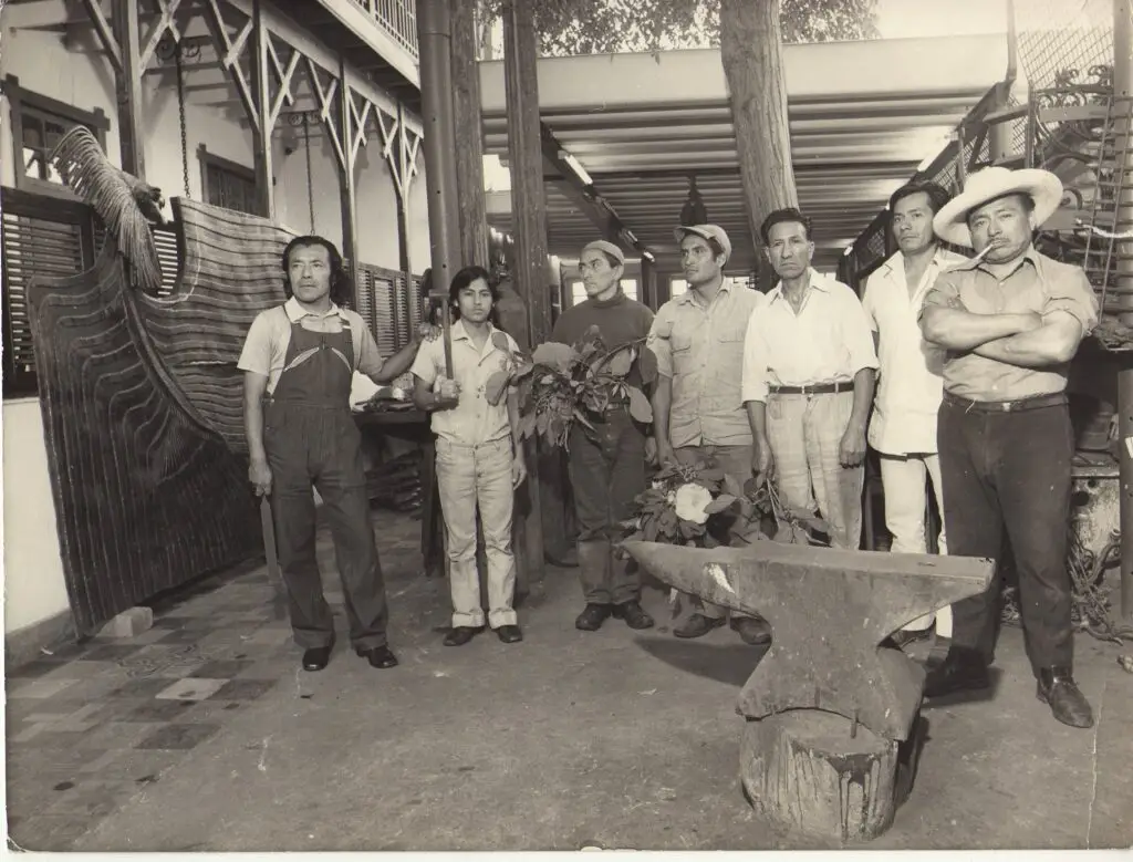 Fotografía en blanco y negro de un grupo de ocho hombres posando en un taller artístico. Al fondo se observa una escultura metálica de gran tamaño en forma de ave. A la izquierda, un hombre con overol sostiene una herramienta, posiblemente el artista. En el centro, algunos hombres sostienen hojas y flores. A la derecha, un hombre con sombrero y pipa cruza los brazos. En primer plano, se ve un yunque sobre un tronco de madera. La arquitectura del lugar muestra columnas, vigas de madera y un segundo piso con barandas, lo que sugiere un espacio de trabajo artesanal o un taller colectivo.