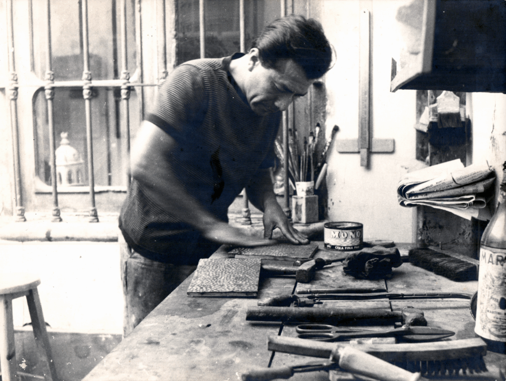 Fotografía en blanco y negro de un hombre trabajando en un taller. Está concentrado puliendo o encerando una superficie sobre una mesa de trabajo cubierta con herramientas. En primer plano hay martillos, limas, brochas y una lata de cera etiquetada “Mono”. Al fondo se aprecian pinceles, papeles enrollados y una botella etiquetada “Mar”, probablemente materiales de arte. A través de la ventana con barrotes de hierro, se distingue la cúpula de un edificio, posiblemente una iglesia. La escena transmite concentración y destreza manual en un ambiente artístico y artesanal.